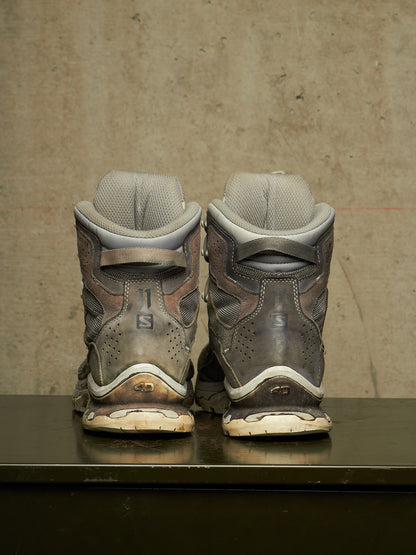 A pair of used Salomon snowboard boots shown from the back, positioned side-by-side on a dark reflective surface against a textured concrete wall.