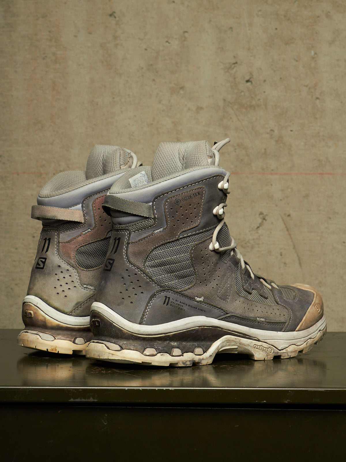 A pair of rugged gray and brown hiking boots with white soles, displayed on a dark reflective surface against a textured concrete wall.