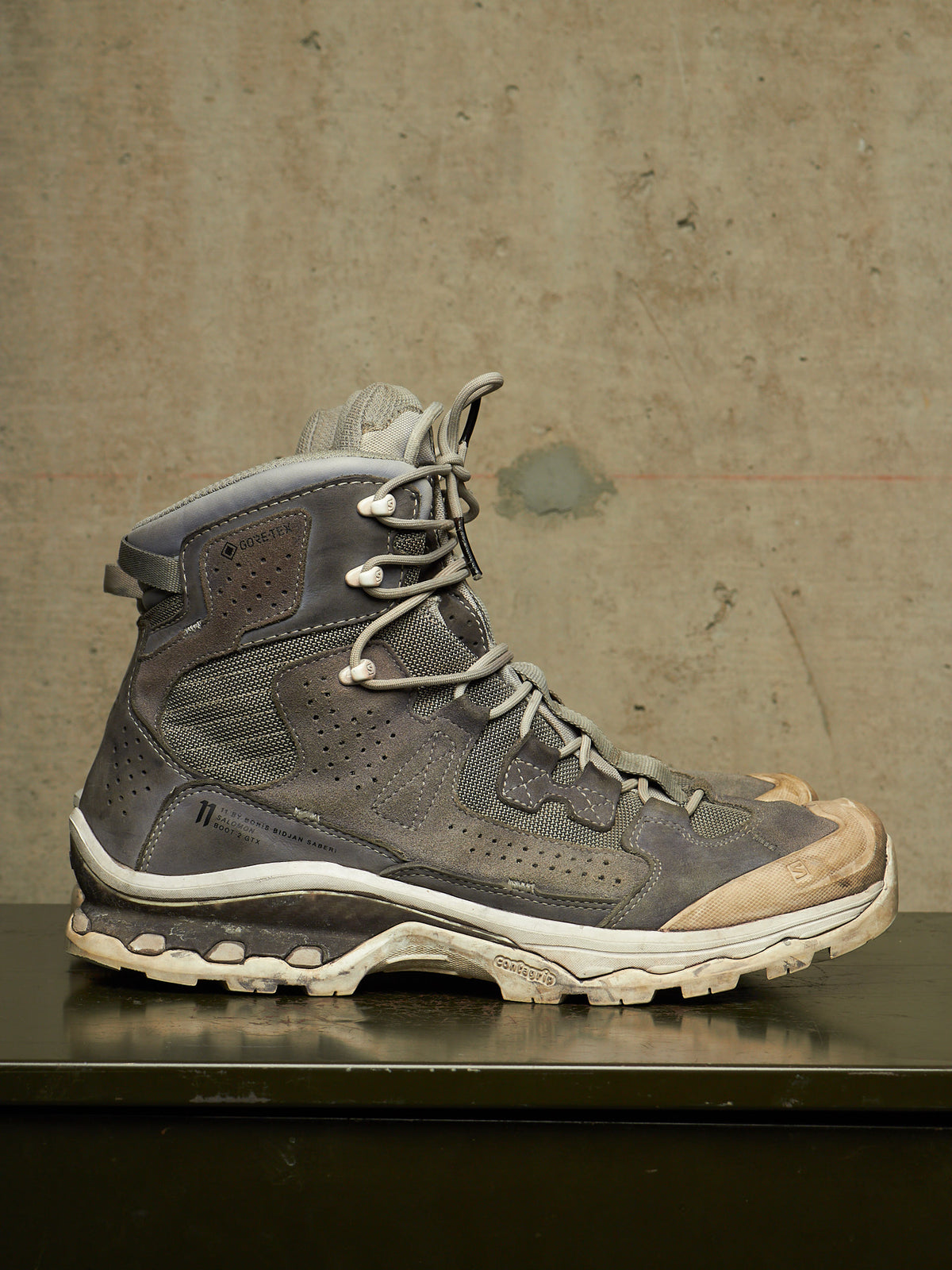 A single high-top hiking boot in distressed gray and brown suede/mesh with white laces and a rugged off-white sole, displayed on a dark surface against a textured concrete wall.