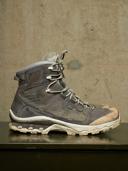 A single high-top hiking boot in distressed gray and brown suede/mesh with white laces and a rugged off-white sole, displayed on a dark surface against a textured concrete wall.