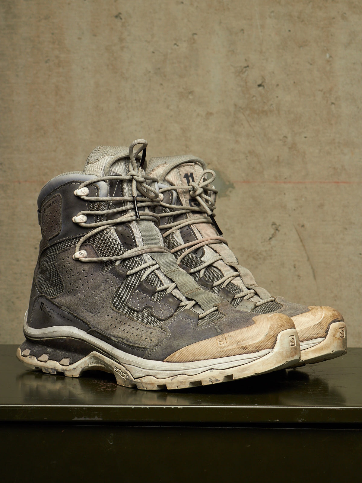 A pair of rugged high-top hiking boots in distressed gray and beige suede/mesh, resting on a dark reflective surface against a textured concrete wall.