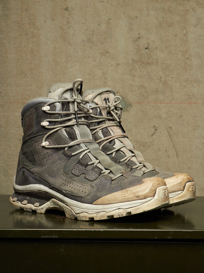 A pair of rugged high-top hiking boots in distressed gray and beige suede/mesh, resting on a dark reflective surface against a textured concrete wall.