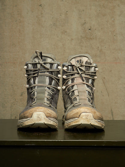 A pair of worn Salomon hiking boots in gray and brown tones, positioned side-by-side on a dark surface against a textured concrete wall.