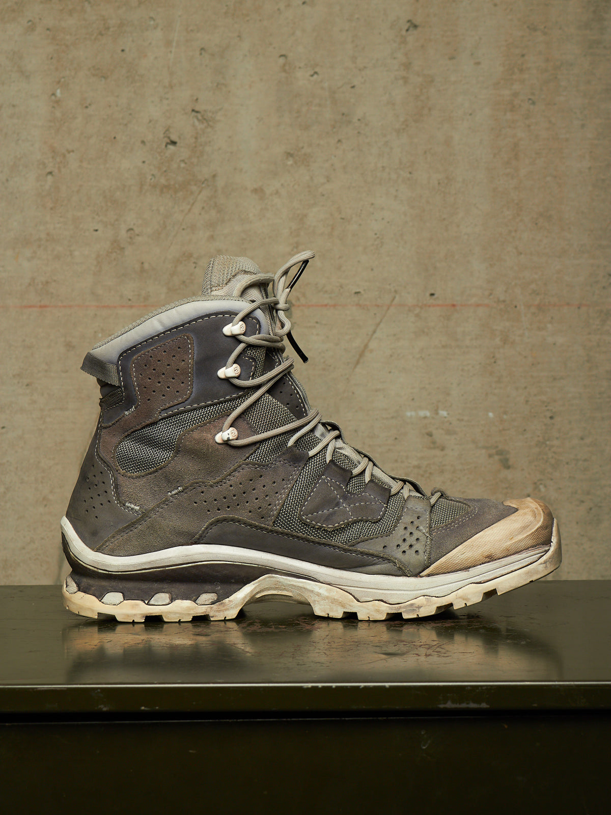 A single high-top lace-up hiking boot in dark gray and brown suede/mesh with a beige-tan toe cap and off-white sole, displayed on a dark reflective surface against a textured concrete wall.