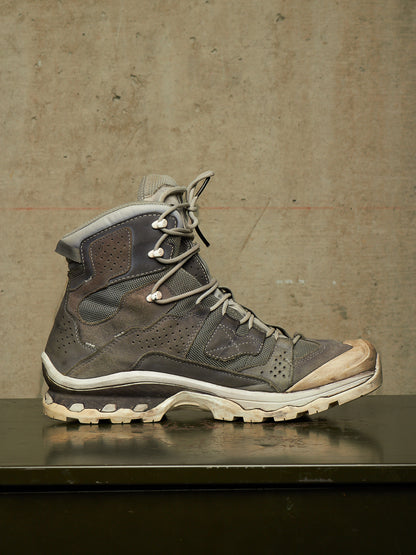 A single high-top lace-up hiking boot in dark gray and brown suede/mesh with a beige-tan toe cap and off-white sole, displayed on a dark reflective surface against a textured concrete wall.