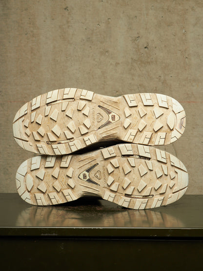 The soles of a pair of rugged outdoor boots, showing deep lugs and tread patterns in a light beige rubber, resting on a dark reflective surface against a textured concrete wall.