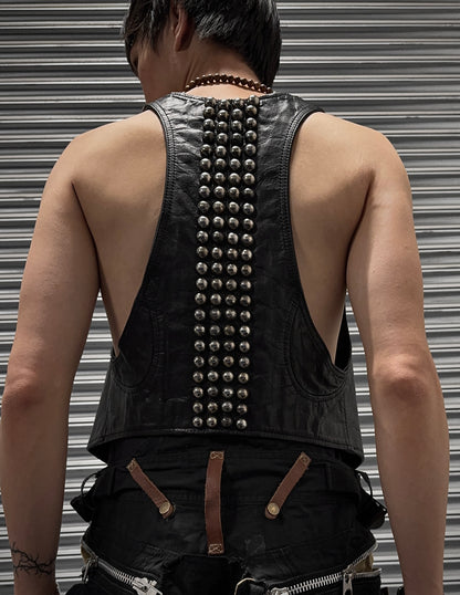 A slim, medium-built person with short dark hair is seen from behind wearing the L.G.B Early 00s Calfskin Leather Studded Mini-vest by L.G.B, styled with pants featuring brown straps, zippers, and subtle discolouration near a metal shutter.