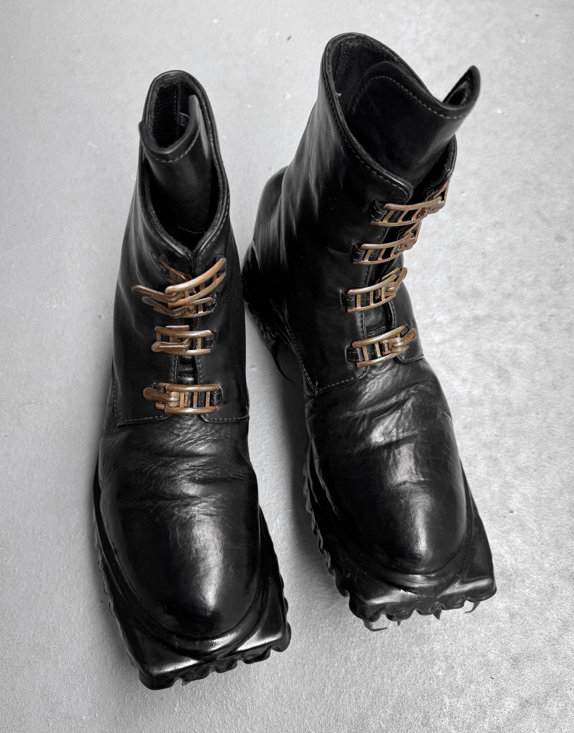 A pair of brand new Carol Christian Poell ‘AM/2759SP-IN CORSS-PTC’ kangaroo leather buckle drip boots in black, size 8, with chunky textured soles and metallic clasps, displayed on a light gray surface.