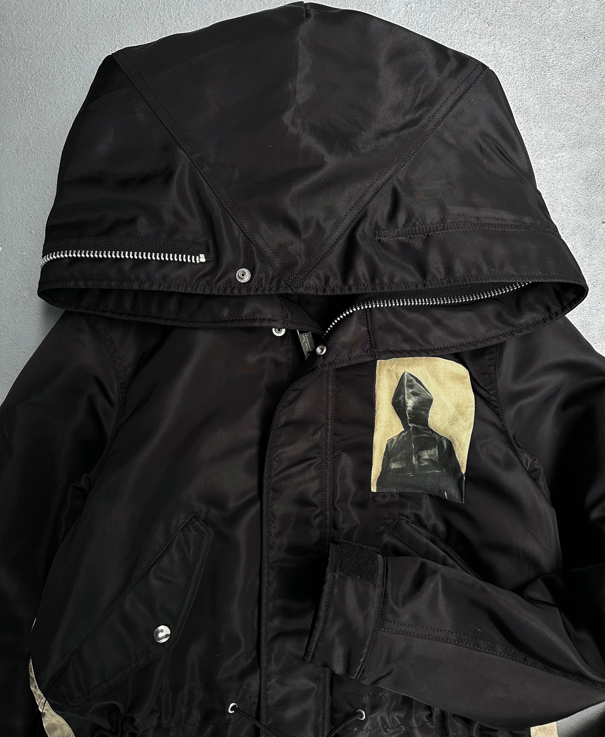 The UNDERCOVER AW16 ‘Borremans’ Full-zip Fishtail Nylon Parka by Undercover is a black hooded jacket with silver snaps and a zipper, featuring a chest patch of a hooded figure. Designed by Jun Takahashi, its pictured on a gray surface.
