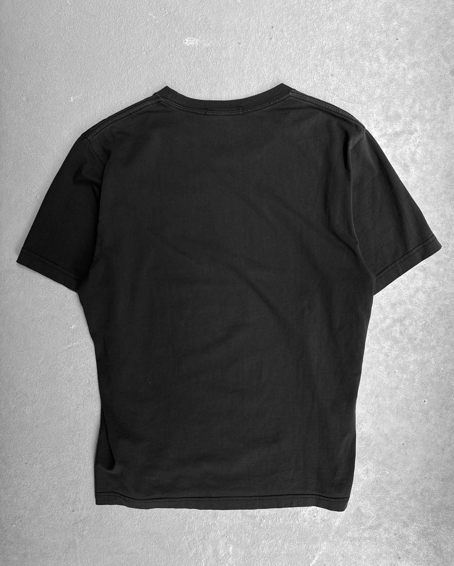 The Undercover SS16 ‘Gilapple’ Graphic Printed T-shirt, a black short-sleeve piece by Undercover, is displayed flat on a light-gray textured surface with its back facing up, highlighting its streetwear appeal.