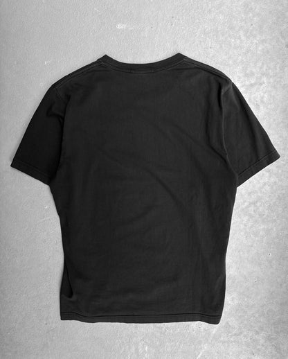 The Undercover SS16 ‘Gilapple’ Graphic Printed T-shirt, a black short-sleeve piece by Undercover, is displayed flat on a light-gray textured surface with its back facing up, highlighting its streetwear appeal.