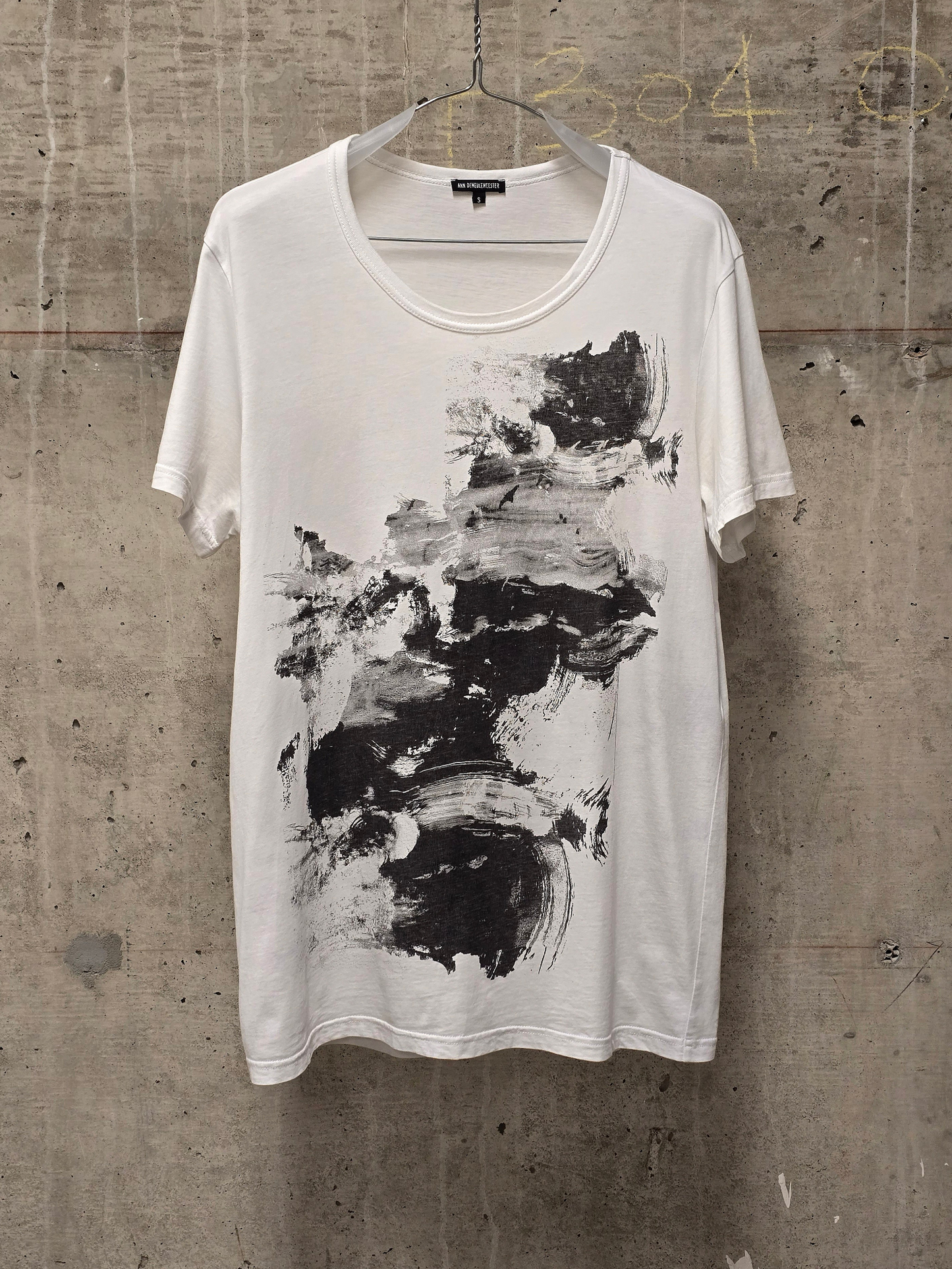 White t-shirt with black abstract design on a hanger against a textured wall.