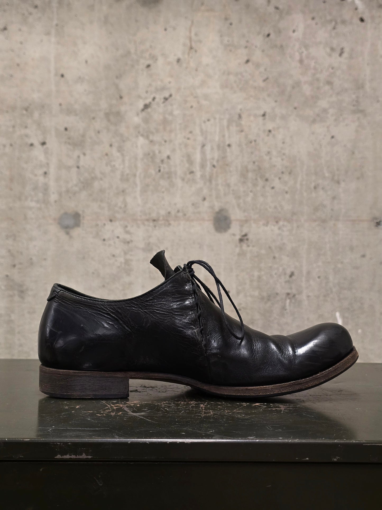 Black leather shoe on a dark surface with a concrete wall background