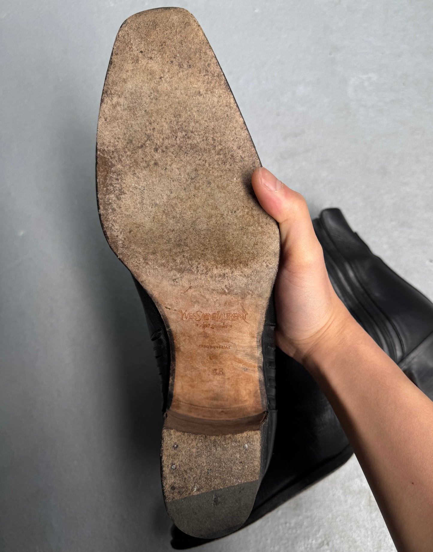 A hand holds up a black leather Saint Laurent Paris YSL Rive Gauche SS04 by Tom Ford 40mm Heel ‘Johnny’ Boot, revealing the worn sole with scuff marks, nails near the heel, and faint branding on the wooden section.