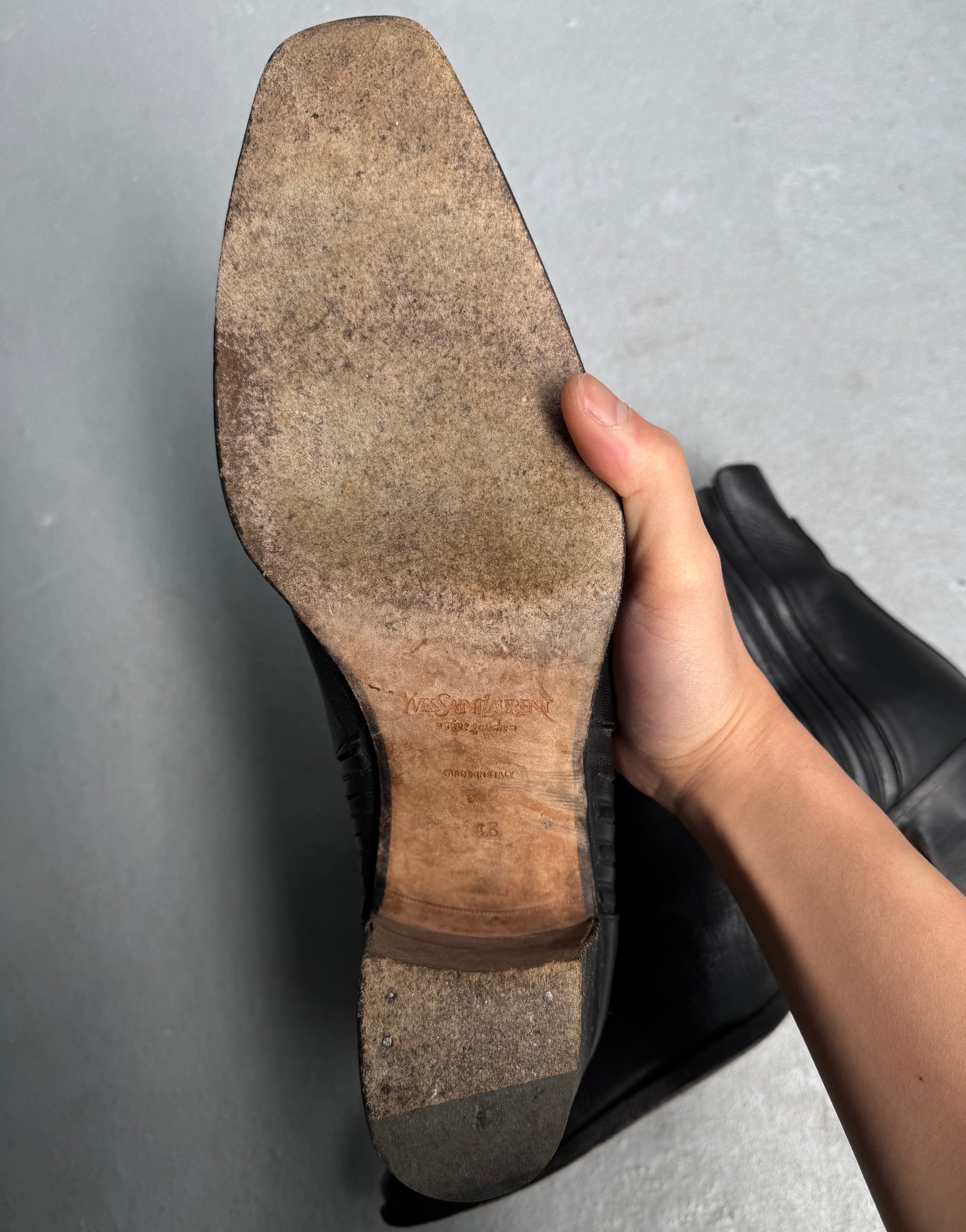 A hand holds up a black leather Saint Laurent Paris YSL Rive Gauche SS04 by Tom Ford 40mm Heel ‘Johnny’ Boot, revealing the worn sole with scuff marks, nails near the heel, and faint branding on the wooden section.