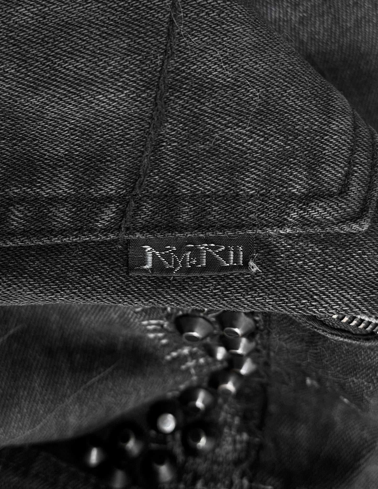 A close-up of KMRii Early 00s Goatskin Patches Studded Crush Flare-zip Denim in Size 2 shows black denim with a small black NMRIK label, subtle fading, and black beads partially visible below the fabric.