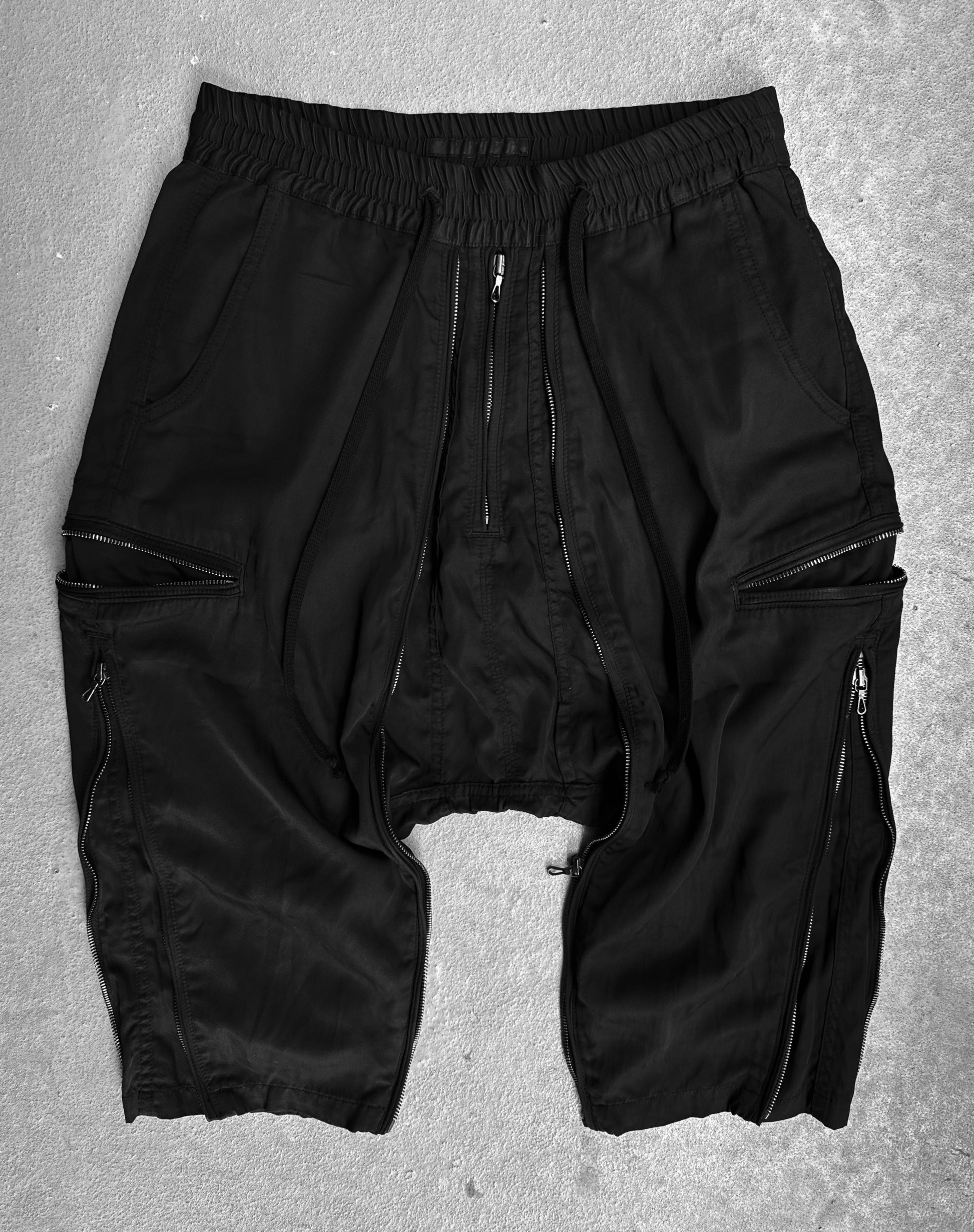 DRIEW Julius_7 SS10 “neurbanvolker;” Convertible Multi-zip Cotton Twill Shorts in black, flawless condition. Drop-crotch design with multiple zippers and pockets. Fits US 29-32 waist (size 2). Laid flat on a textured gray surface.