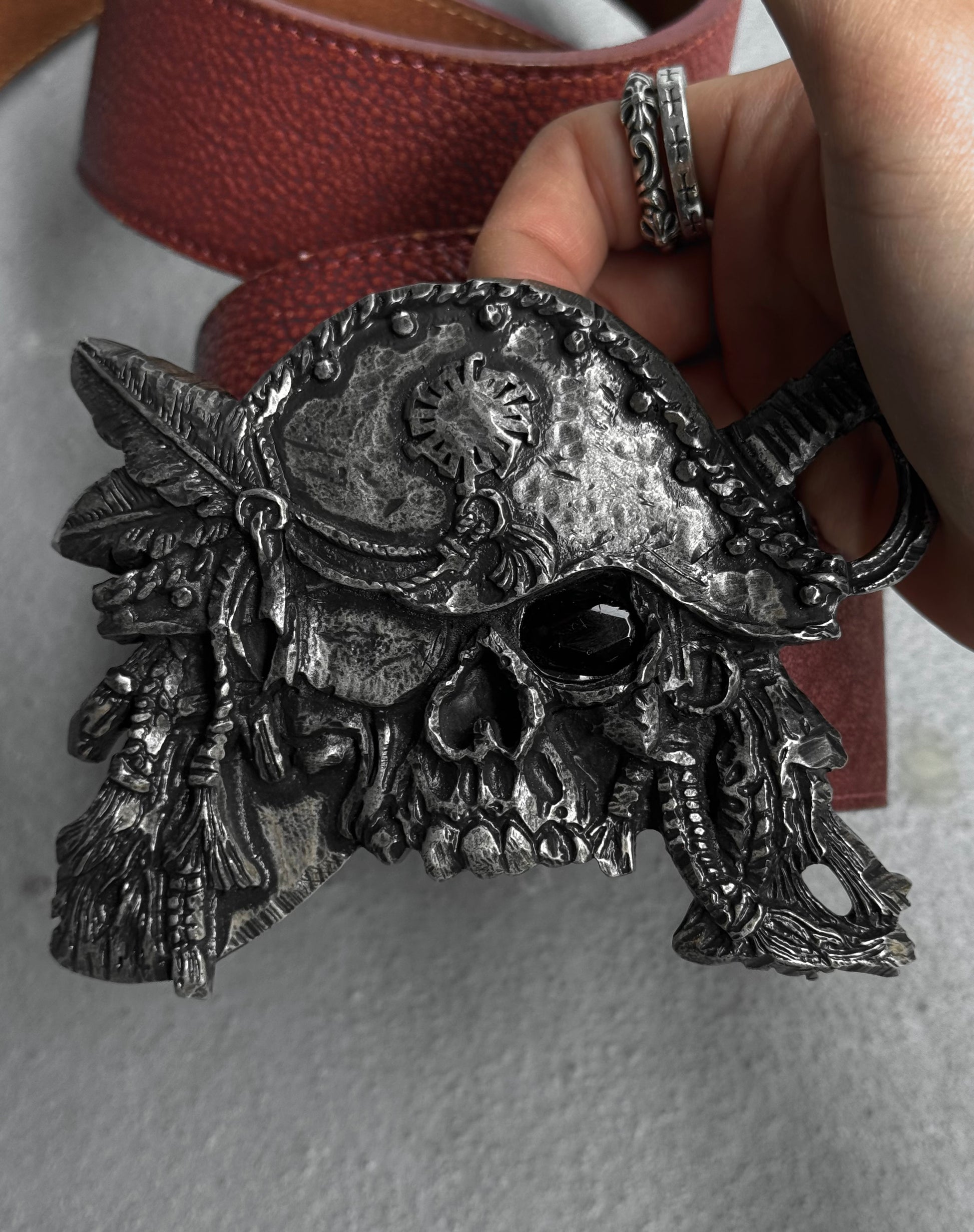 A hand holds the Tornado Mart Early 00s Pirate Silver Buckle Leather Belt—featuring a skull-shaped metallic buckle with ornate hat and red leather strap. Size M fits US 32-36 waist and is in flawless condition.