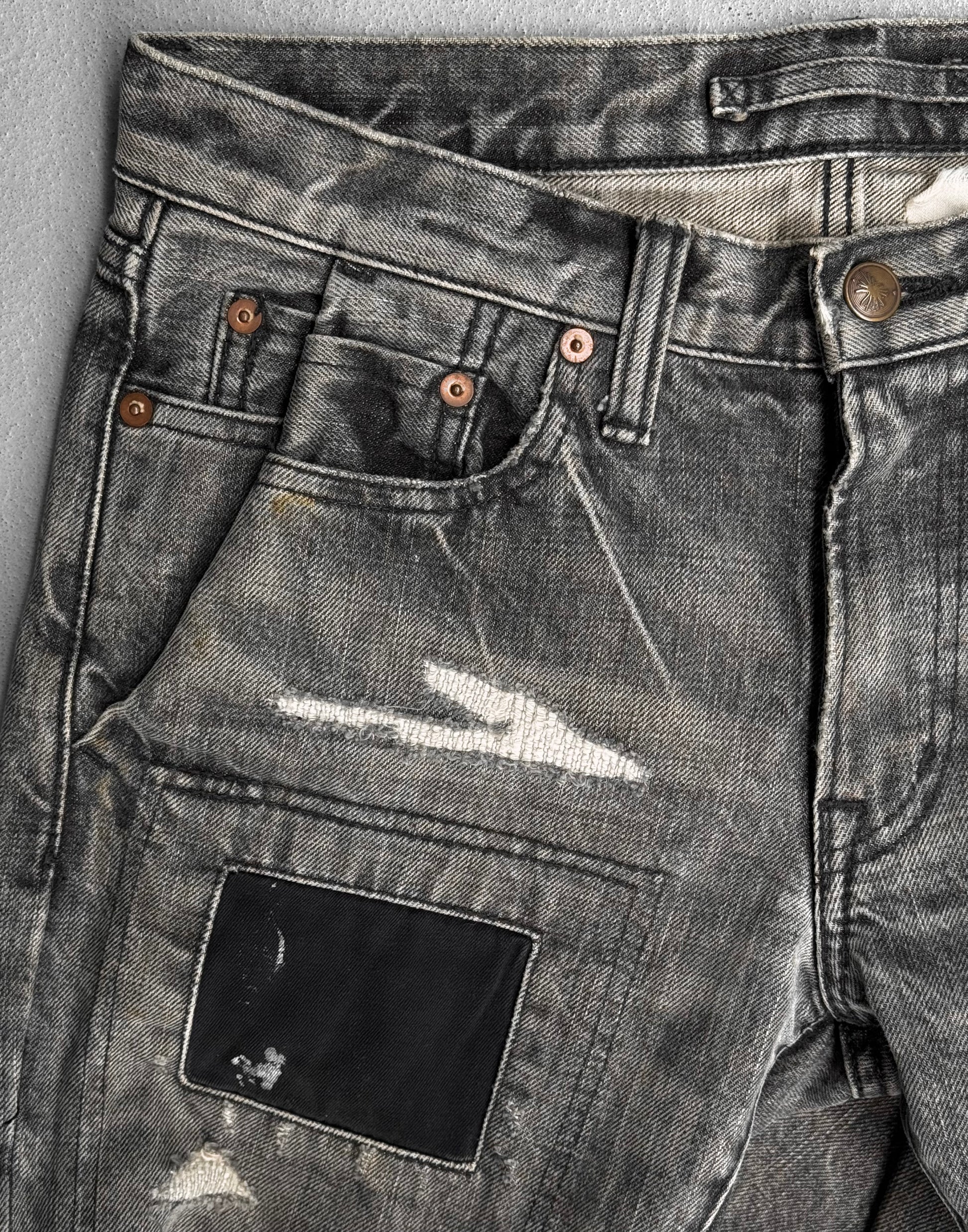 A close-up of Mihara Yasuhiro AW10 Double-pockets Repair Patchwork Selvedge Denim shows distressed gray jeans with fading, rips, frayed edges, copper rivets, and a dark repair patch—reflecting Mihara Yasuhiros signature streetwear edge.