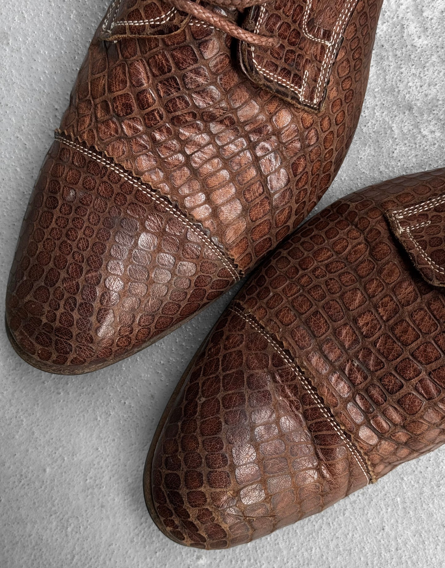 Close-up of Mihara Yasuhiro Early 00s Python Embossed Cow Leather Pointy Toe Boots, featuring detailed stitching and laces on a light gray textured surface—an elegant nod to avant-garde Japanese design.