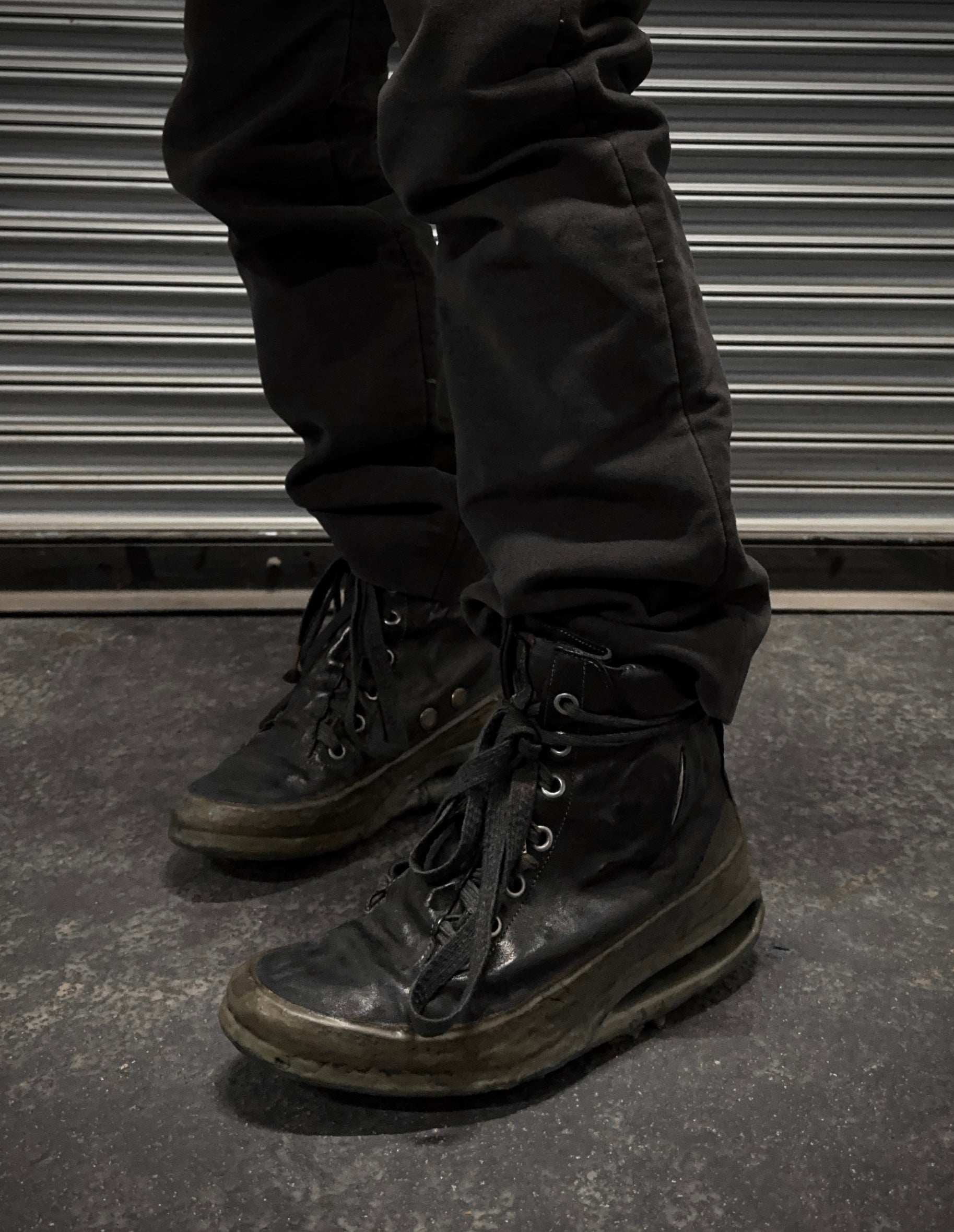 A person in black pants and DRIEW Carol Christian Poell ‘AM/2685P ROOMS-PTC/12’ object-dyed kangaroo leather titanium prosthetic U-sole drip sneakers, size 6 with light wear and untied laces, stands on a dark concrete floor by a ribbed metallic shutter.