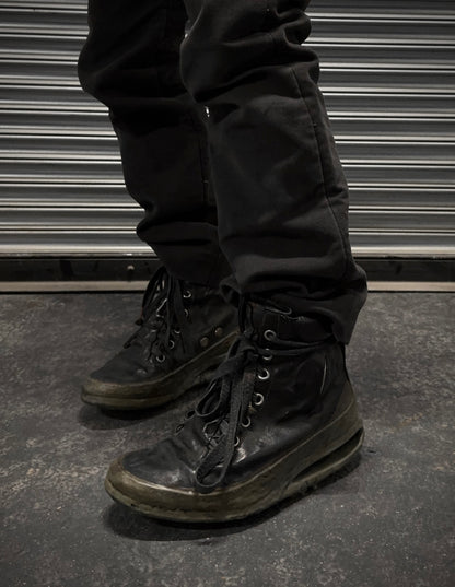 A person in black pants and DRIEW Carol Christian Poell ‘AM/2685P ROOMS-PTC/12’ object-dyed kangaroo leather titanium prosthetic U-sole drip sneakers, size 6 with light wear and untied laces, stands on a dark concrete floor by a ribbed metallic shutter.