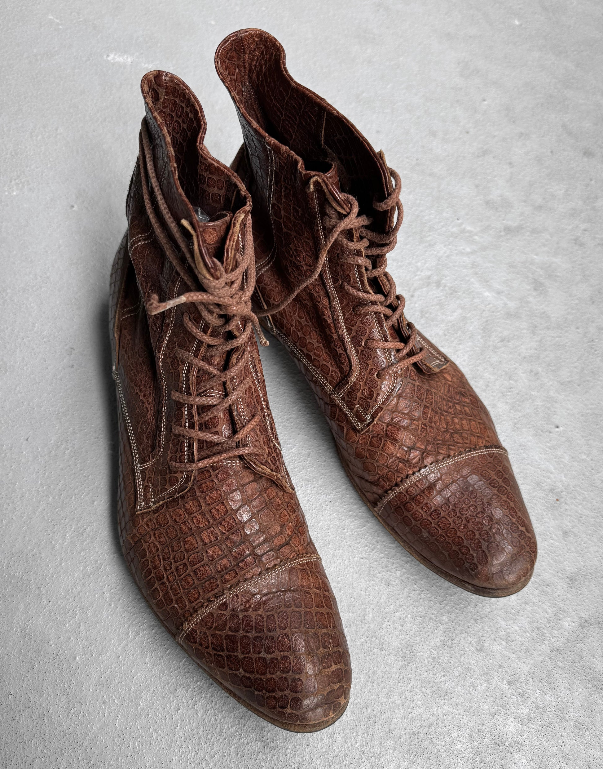 A pair of Mihara Yasuhiro Early 00s Python Embossed Cow Leather Pointy Toe Boots, featuring a brown reptile-textured surface and lace-up design, are placed on a light gray floor.