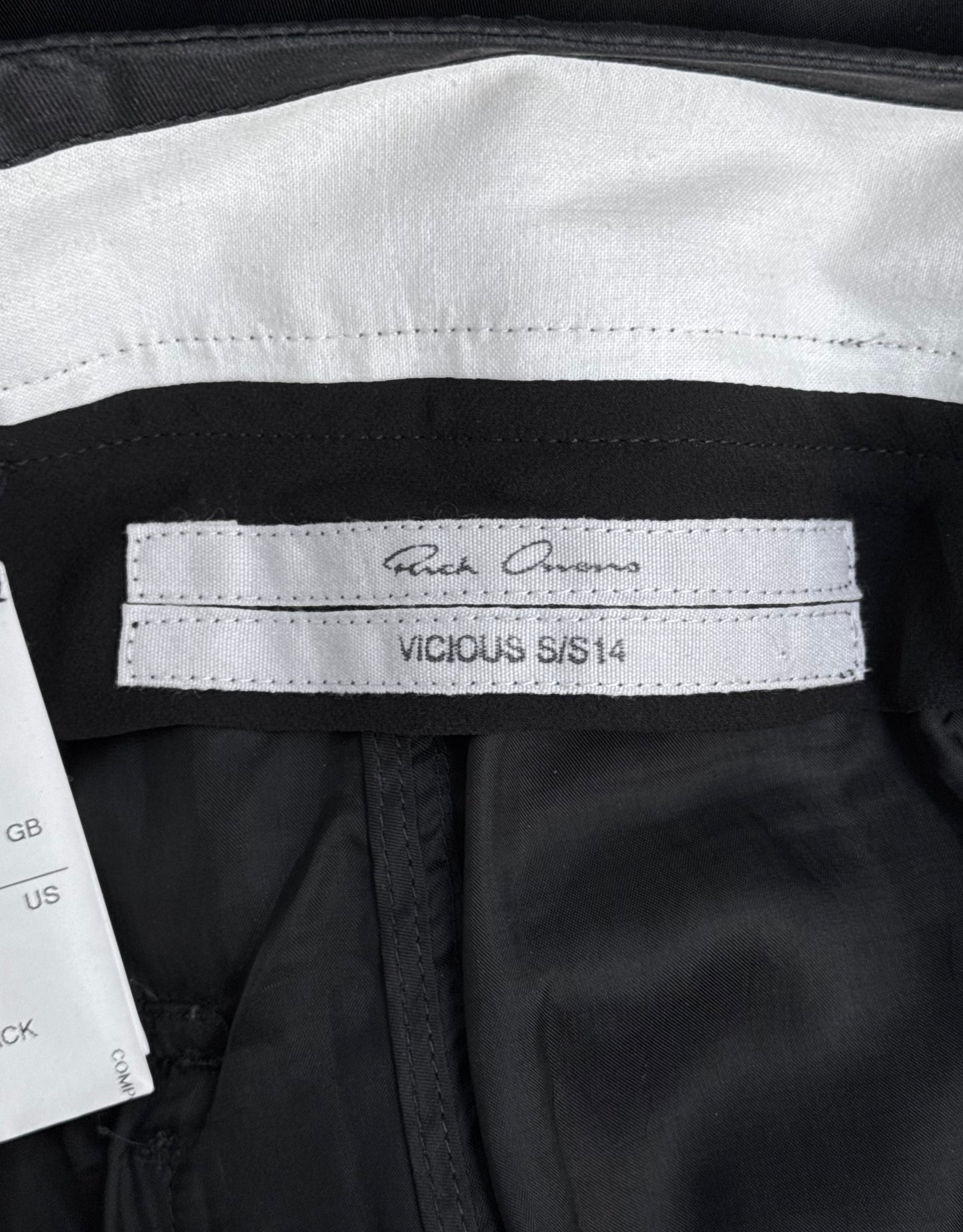 A close-up of a label inside Rick Owens SS14 “VICIOUS” Lightweight Memphis Shorts, featuring a signature-style logo; a white care tag with size info appears partially visible on the left, reflecting the brands contemporary style.
