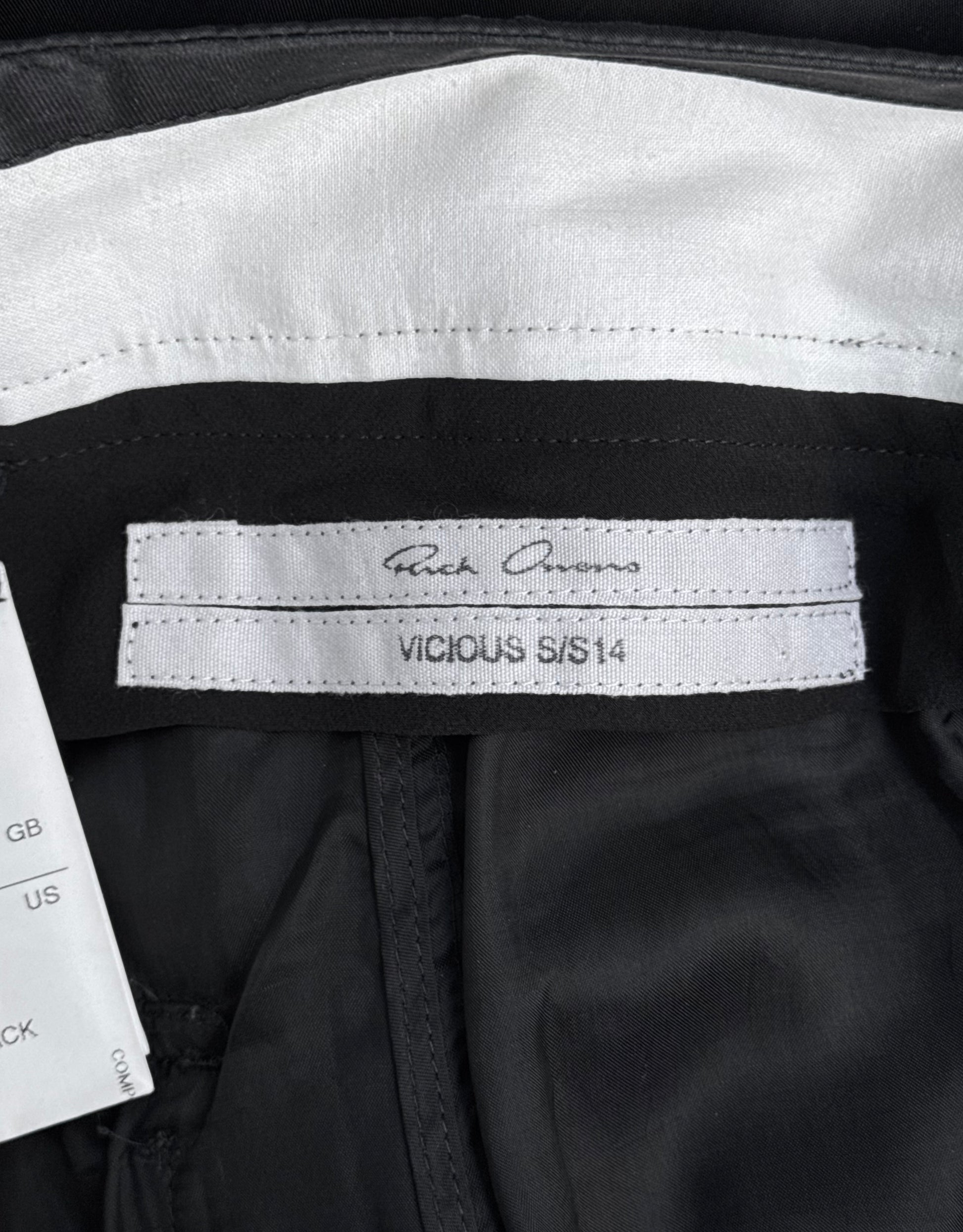 A close-up of a label inside Rick Owens SS14 “VICIOUS” Lightweight Memphis Shorts, featuring a signature-style logo; a white care tag with size info appears partially visible on the left, reflecting the brands contemporary style.