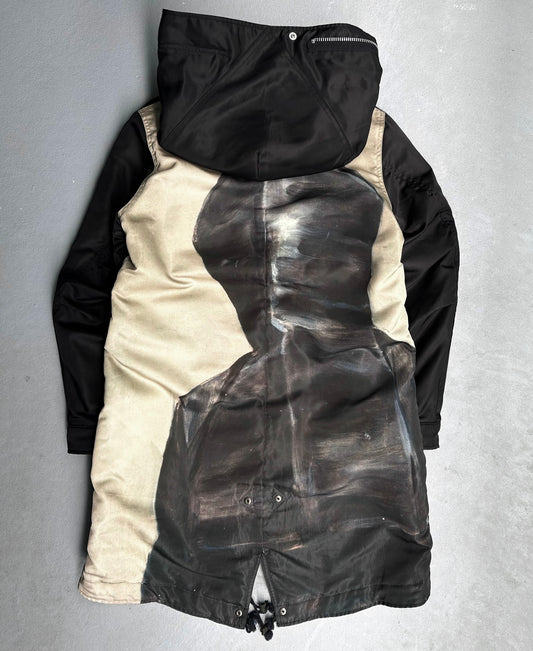 The UNDERCOVER AW16 ‘Borremans’ Full-zip Fishtail Nylon Parka by Undercover features black sleeves and hood, plus a large painted dark silhouette on the tan back panel, shown on a gray surface.