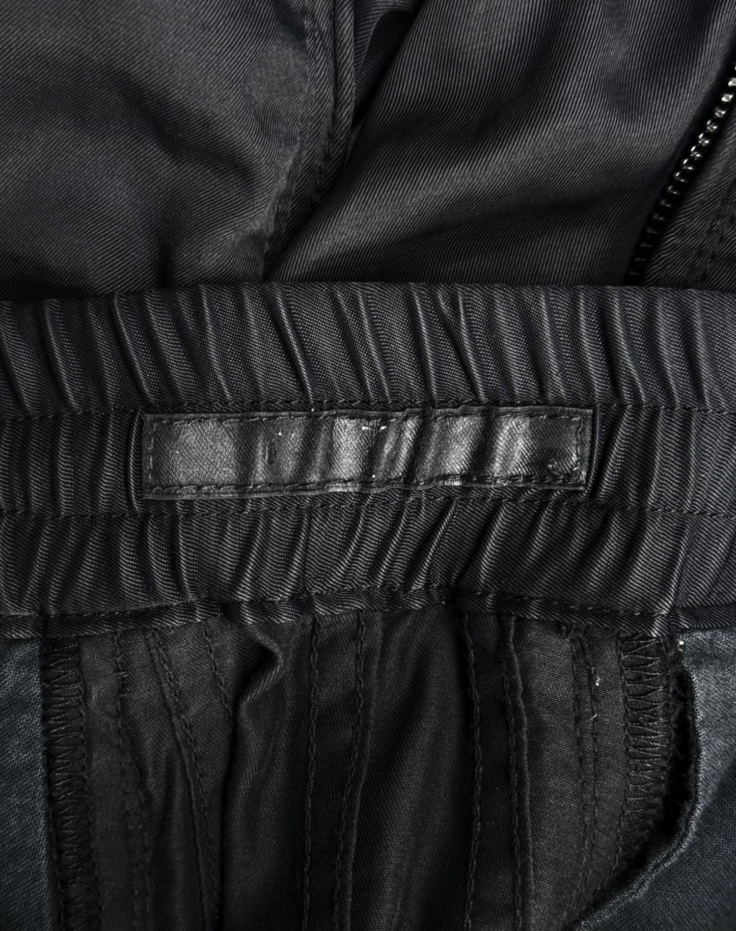 Close-up of the inside waistband of Julius_7 SS10 “neurbanvolker;” Convertible Multi-zip Cotton Twill Shorts by DRIEW in black, showing an empty black leather label, gathered fabric, visible stitching, part of a zipper, fits US 29-32 or size 2.