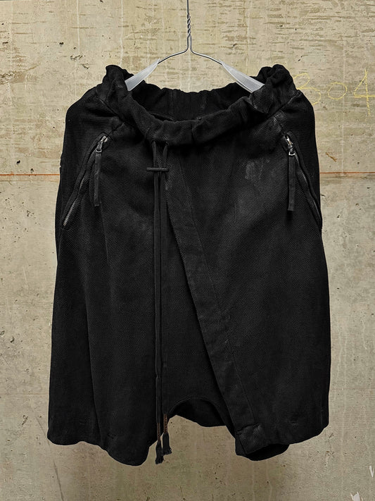 A black asymmetrical wrap skirt with a drawstring waist and zippered side pockets, hanging on a wire hanger against a textured concrete wall.