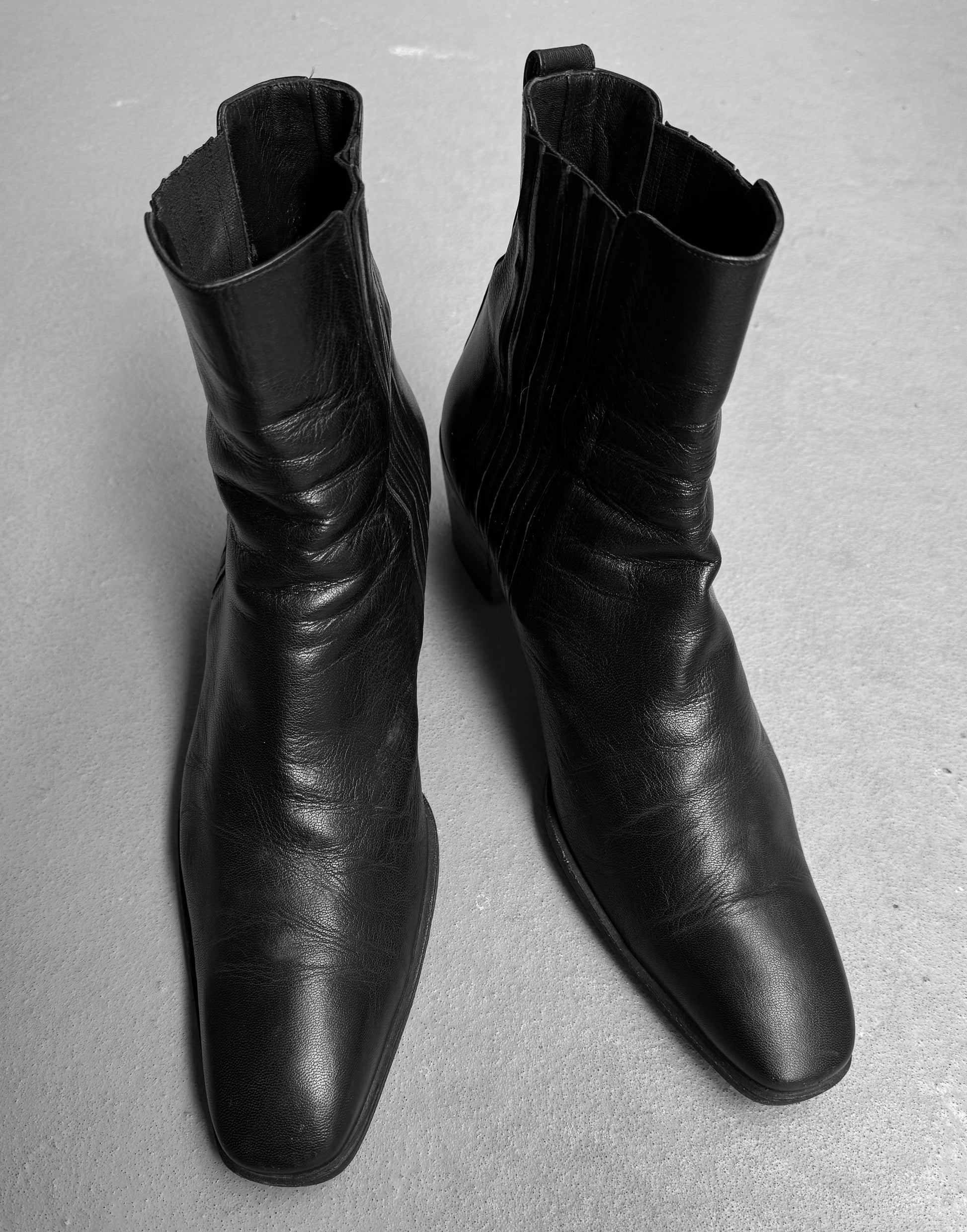 A pair of Saint Laurent Paris YSL Rive Gauche SS04 by Tom Ford 40mm Heel ‘Johnny’ black leather ankle boots with square toes, inspired by Yves Saint Laurent, placed side by side on a light-colored floor.