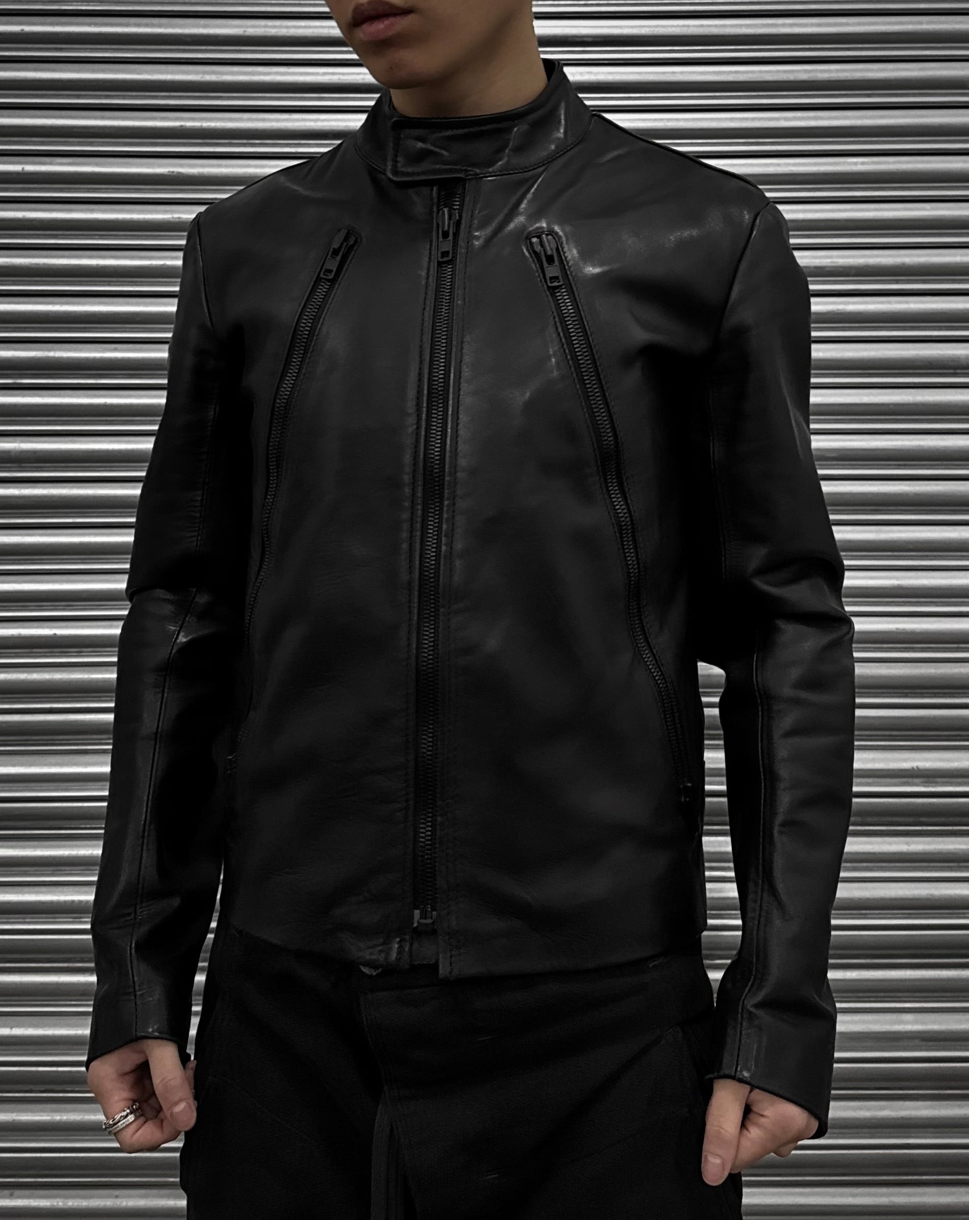 A person models a Maison Martin Margiela AW10-11 Kokone Black Horse Leather 5-zip Jacket (size 44) with vertical zippers, standing before a metallic corrugated shutter. The photo is cropped above the mouth and below the waist.