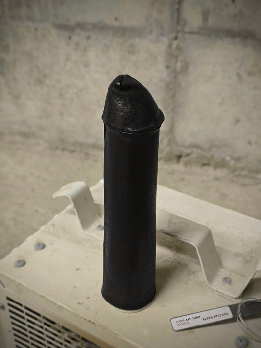 A black silicone adult novelty product, specifically a realistic dildo, standing upright on a beige plastic display stand against a gray concrete block wall.