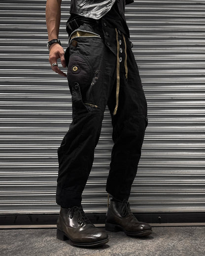 A person stands before a metal shutter wearing L.G.B Early 00s Pilot Cargo Capris (US 32 Waist, used), featuring repair patches, button details, and a belt with metal rings. They pair them with dark boots and several rings and bracelets; torso is partially out of frame.