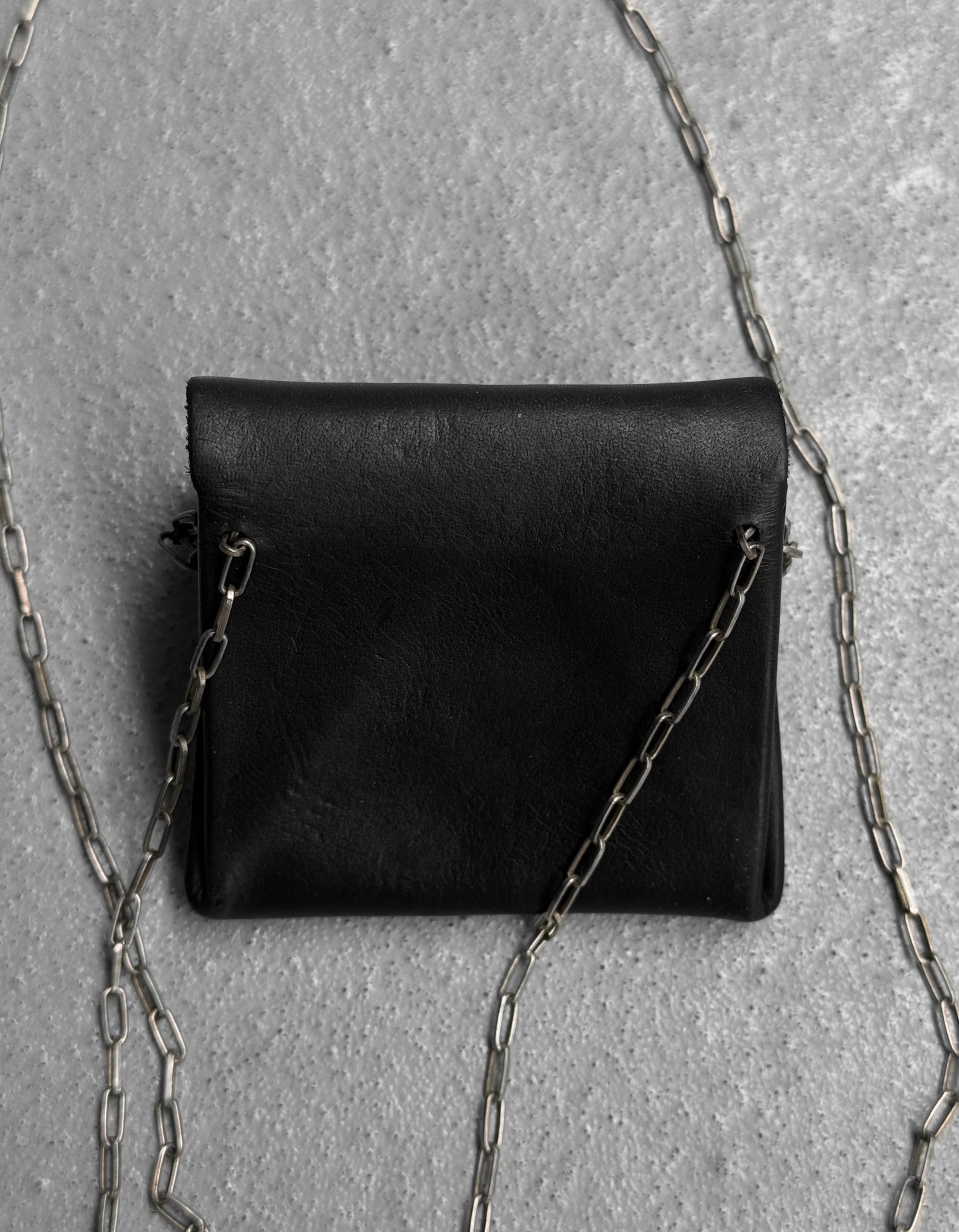 The M.A+ by Maurizio Amadei ‘A-B7201’ Calfskin Leather Mini Accordion Pouch Necklace in black OneSize is displayed on a textured gray surface, its elegant silver chain strap gracefully curved around the pouch.