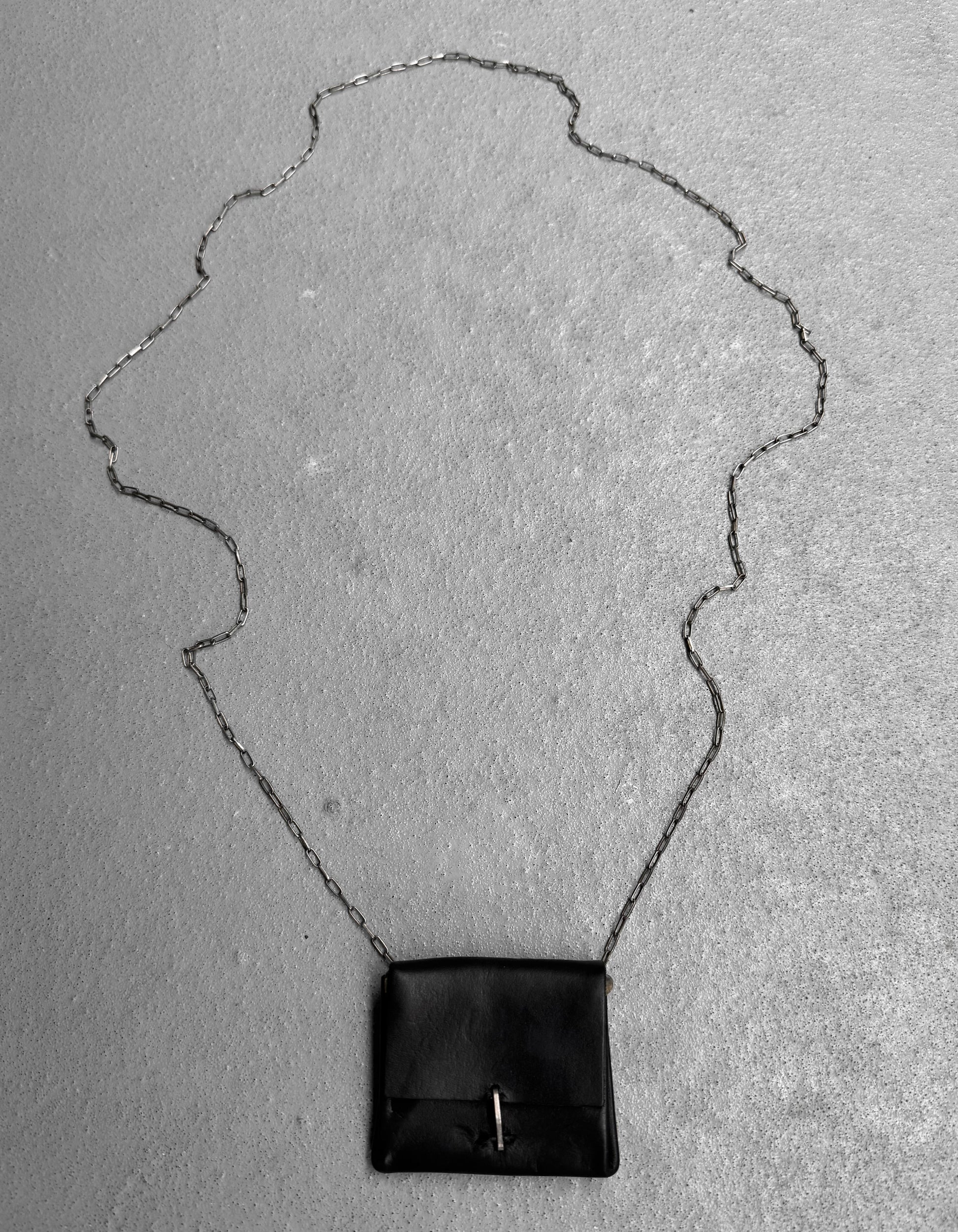 A small black M.A+ by Maurizio Amadei ‘A-B7201’ Calfskin Leather Mini Accordion Pouch Necklace with a long silver chain is displayed on a textured gray surface, its decorative chain forming an elegant shape around the piece.