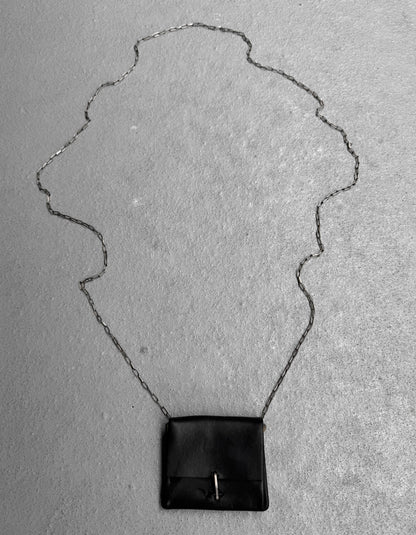 A small black M.A+ by Maurizio Amadei ‘A-B7201’ Calfskin Leather Mini Accordion Pouch Necklace with a long silver chain is displayed on a textured gray surface, its decorative chain forming an elegant shape around the piece.