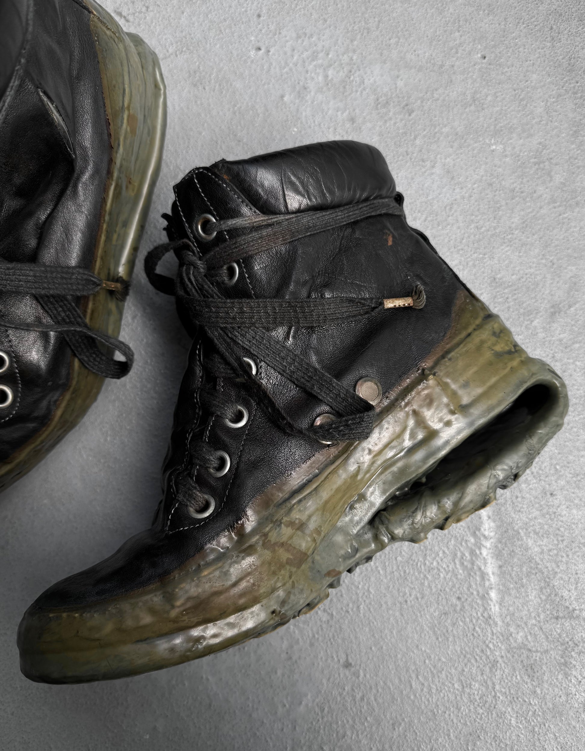 A worn DRIEW Carol Christian Poell ‘AM/2685P ROOMS-PTC/12’ black kangaroo leather sneaker, Size 6, with a cracked, melted titanium U-sole is displayed on a gray surface; part of another similarly damaged sneaker is seen in the corner.