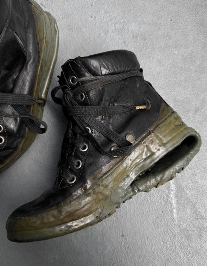 A worn DRIEW Carol Christian Poell ‘AM/2685P ROOMS-PTC/12’ black kangaroo leather sneaker, Size 6, with a cracked, melted titanium U-sole is displayed on a gray surface; part of another similarly damaged sneaker is seen in the corner.