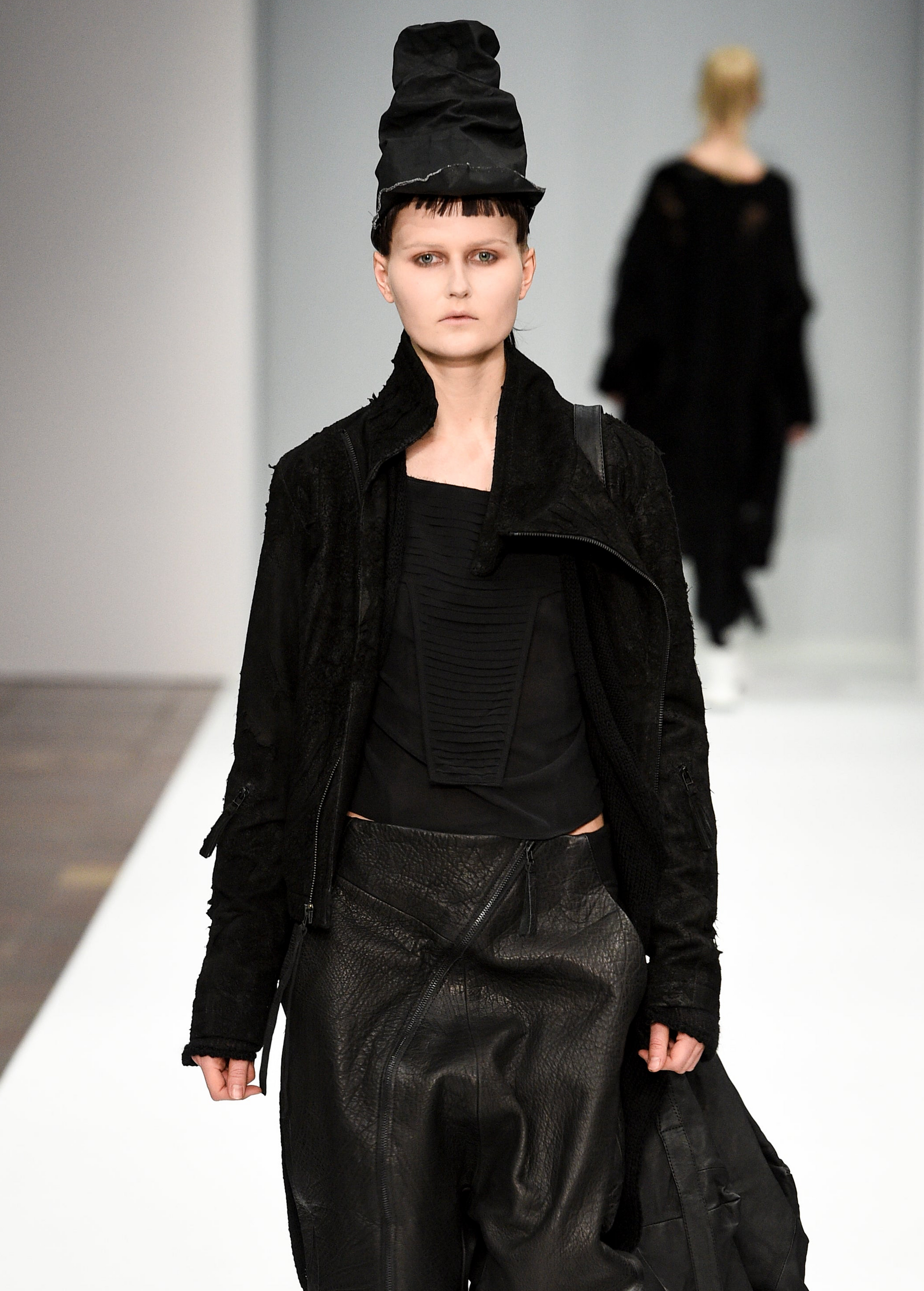 A runway photo of a model wearing a black textured jacket, black pleated top, black wide-leg pants, and a tall black hat, walking on a fashion show catwalk.