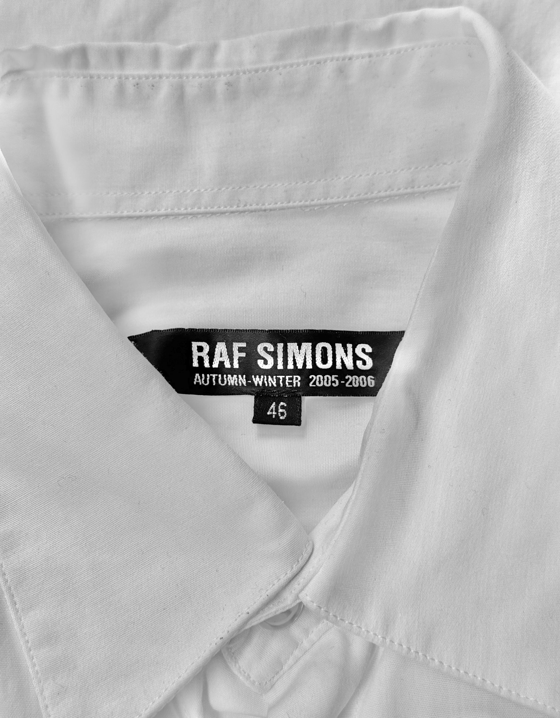 A close-up of the RAF SIMONS AW05-06 “History Of My World” Extended Sleeves Button-up Shirt shows a black label with brand and season details, size 46, reflecting RAF SIMONS impact on contemporary fashion.