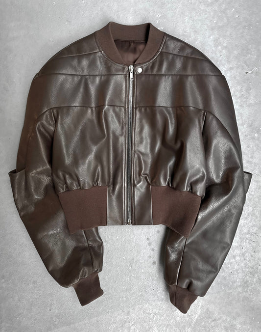 The Rick Owens AW23 LUXOR Girdered Leather Bomber Jacket, in cropped brown calfskin with a front zipper and ribbed collar, cuffs, and waistband, is shown laid flat on a gray surface.