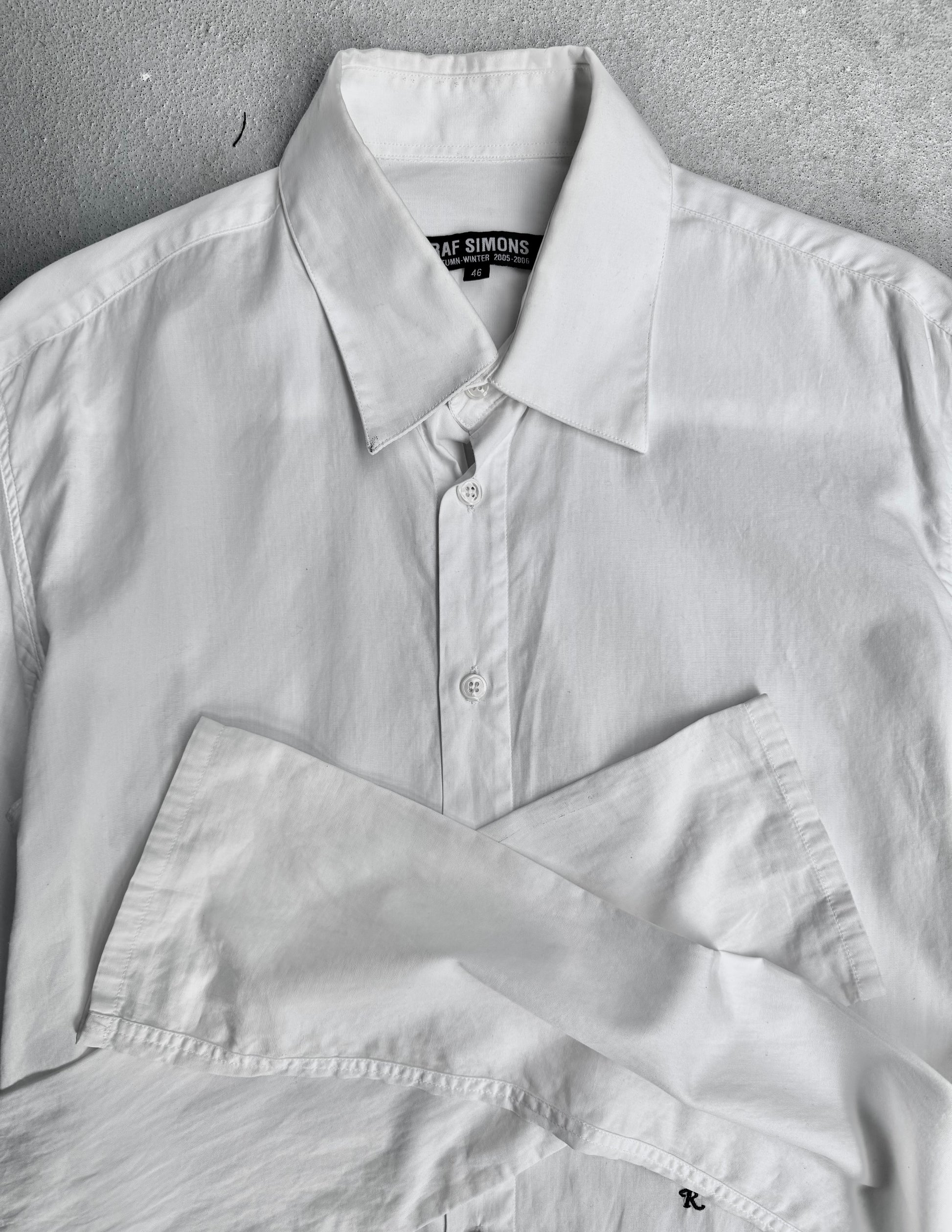 A RAF SIMONS AW05-06 “History Of My World” Extended Sleeves Button-up Shirt in white is neatly folded on a light grey surface, highlighting its collar, buttons, and refined contemporary style.