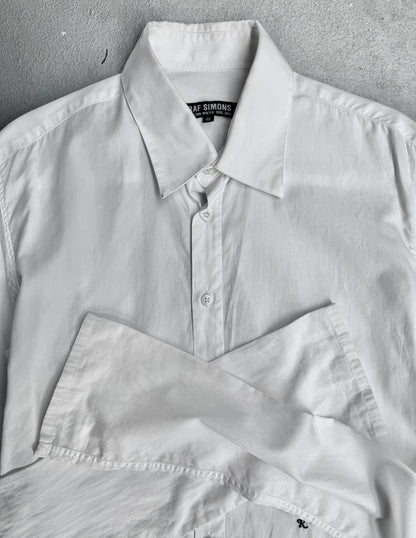 A RAF SIMONS AW05-06 “History Of My World” Extended Sleeves Button-up Shirt in white is neatly folded on a light grey surface, highlighting its collar, buttons, and refined contemporary style.