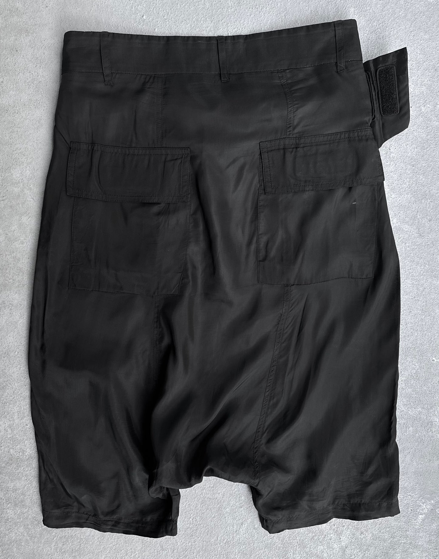 The Rick Owens SS14 “VICIOUS” Lightweight Memphis Shorts, in black, feature two large back flap pockets, an adjustable waistband strap, and a slightly wrinkled texture, shown laid flat on a light gray surface.