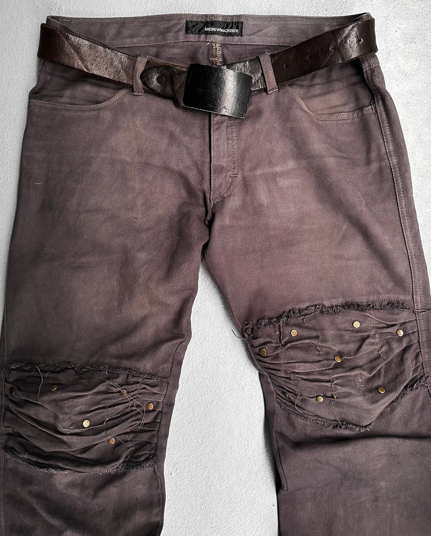 ANDREW MACKENZIE Early 00s Stud Darted Knee Denim: Distressed dark brown jeans with large knee rips, folded knee detailing, and metal rivets, shown flat with a worn leather belt.