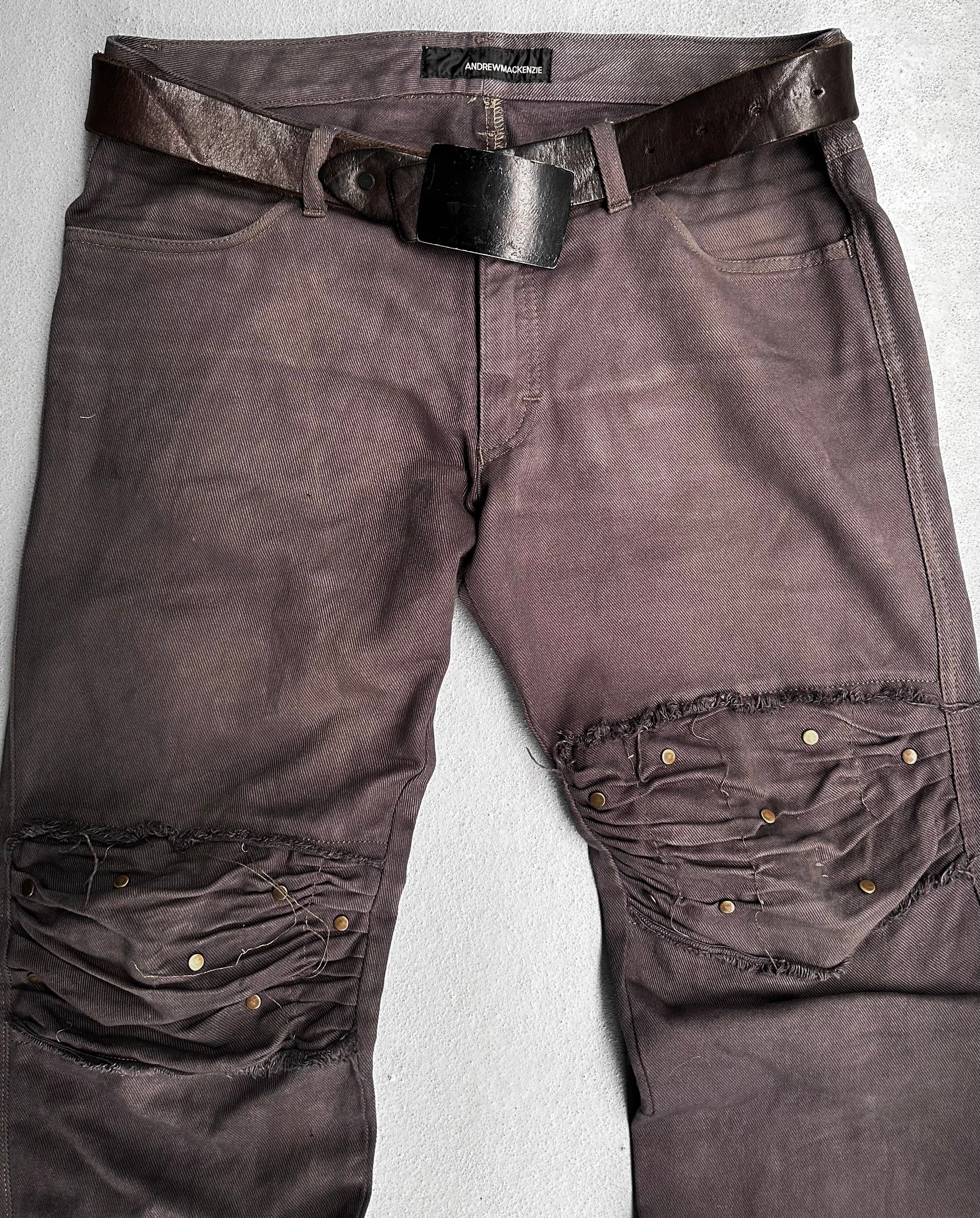 ANDREW MACKENZIE Early 00s Stud Darted Knee Denim: Distressed dark brown jeans with large knee rips, folded knee detailing, and metal rivets, shown flat with a worn leather belt.