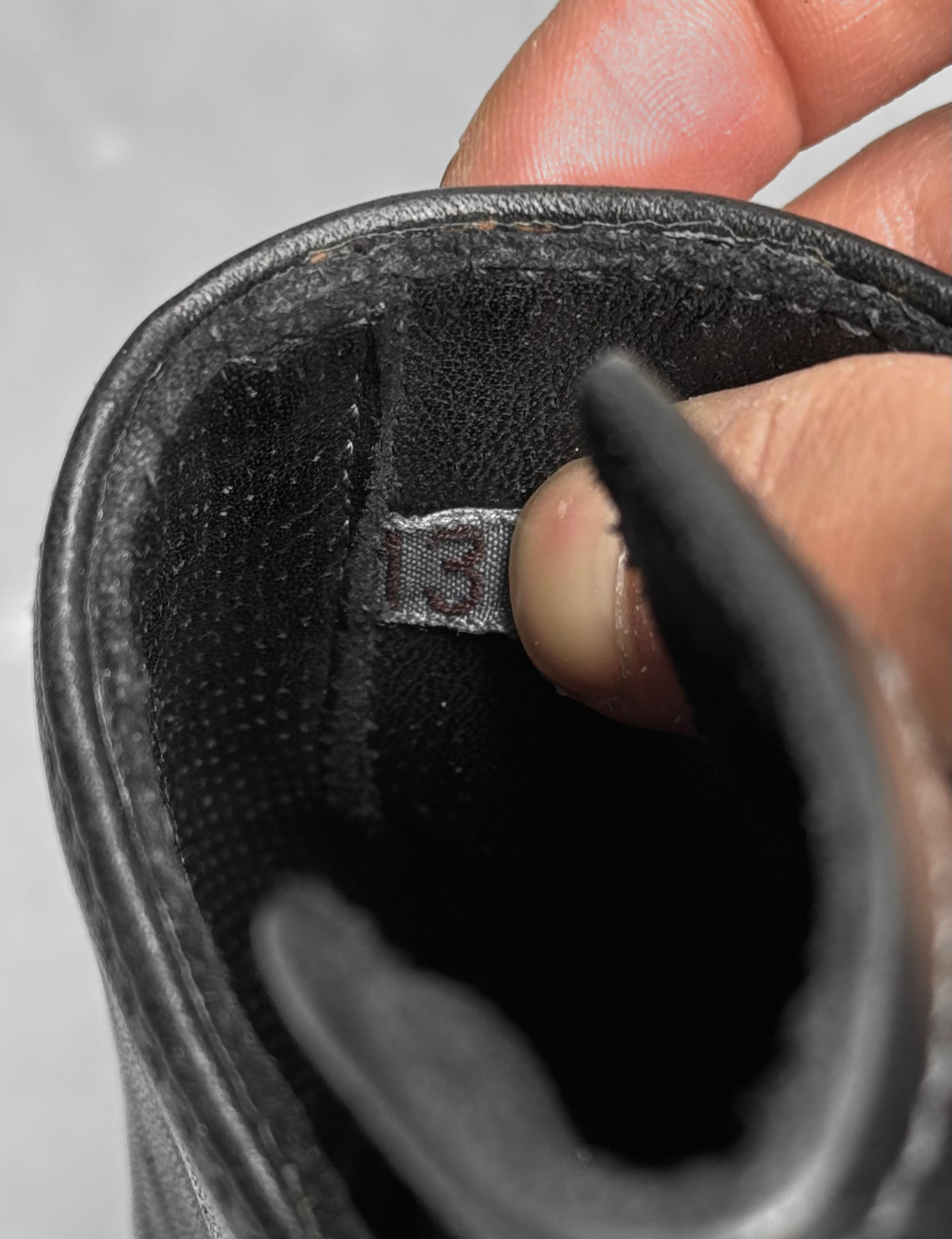 A close-up of a hand holding open the tongue of a new Carol Christian Poell ‘AM/2759SP-IN CORSS-PTC’ Kangaroo Leather Buckle Drip Boot, revealing a fabric tag with the number 13.